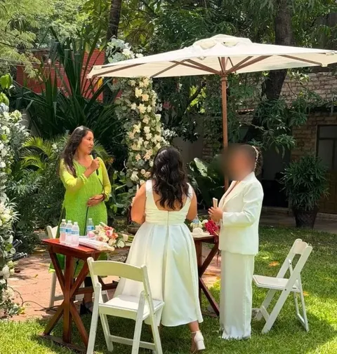 Tanis, de verde y con micrófono, oficia una boda en un jardín; la pareja la escucha de espaldas