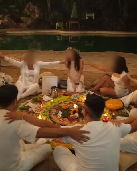 Un Cumple Sol de noche en la arena: el círculo, de blanco, alrededor de un mandala de flores y velas, y al centro quien cumple años, con su corona de flores